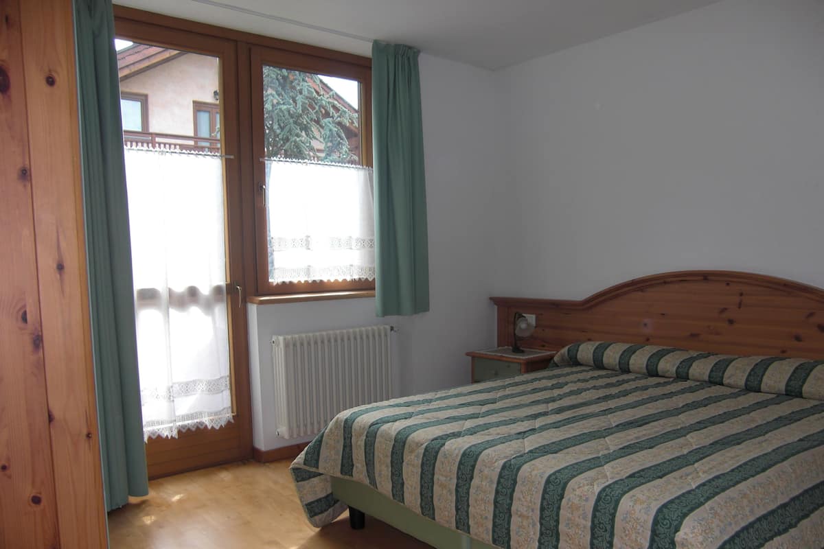 A bedroom with a wooden bed, a nightstand, a window with curtains, and a radiator.