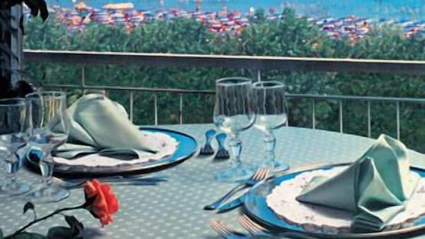 Mesa para dos con vista al océano y sombrillas de playa al fondo.
