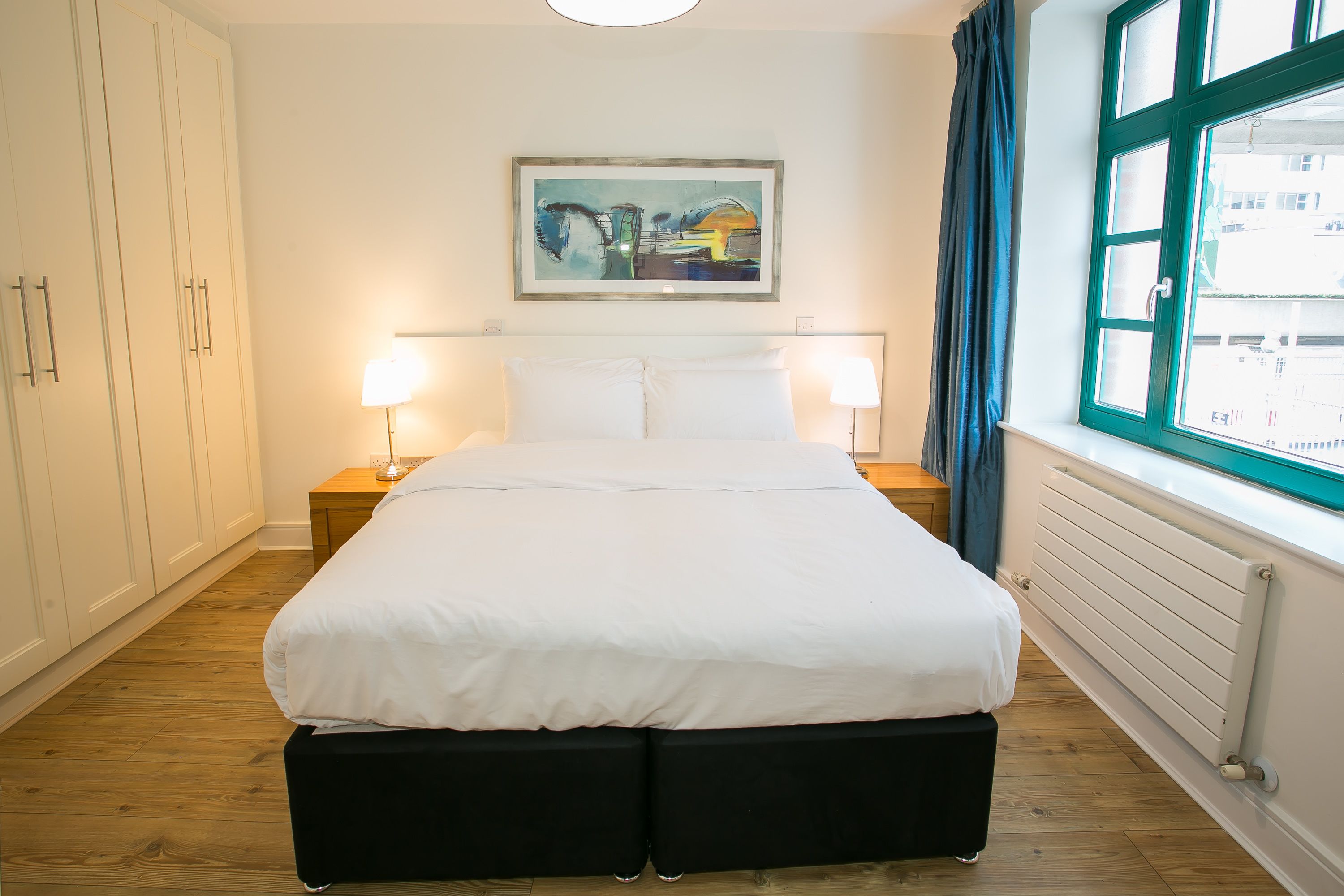 A neatly made bed with white linens, two bedside tables with lamps, a large abstract painting above the bed, and a window with blue curtains.