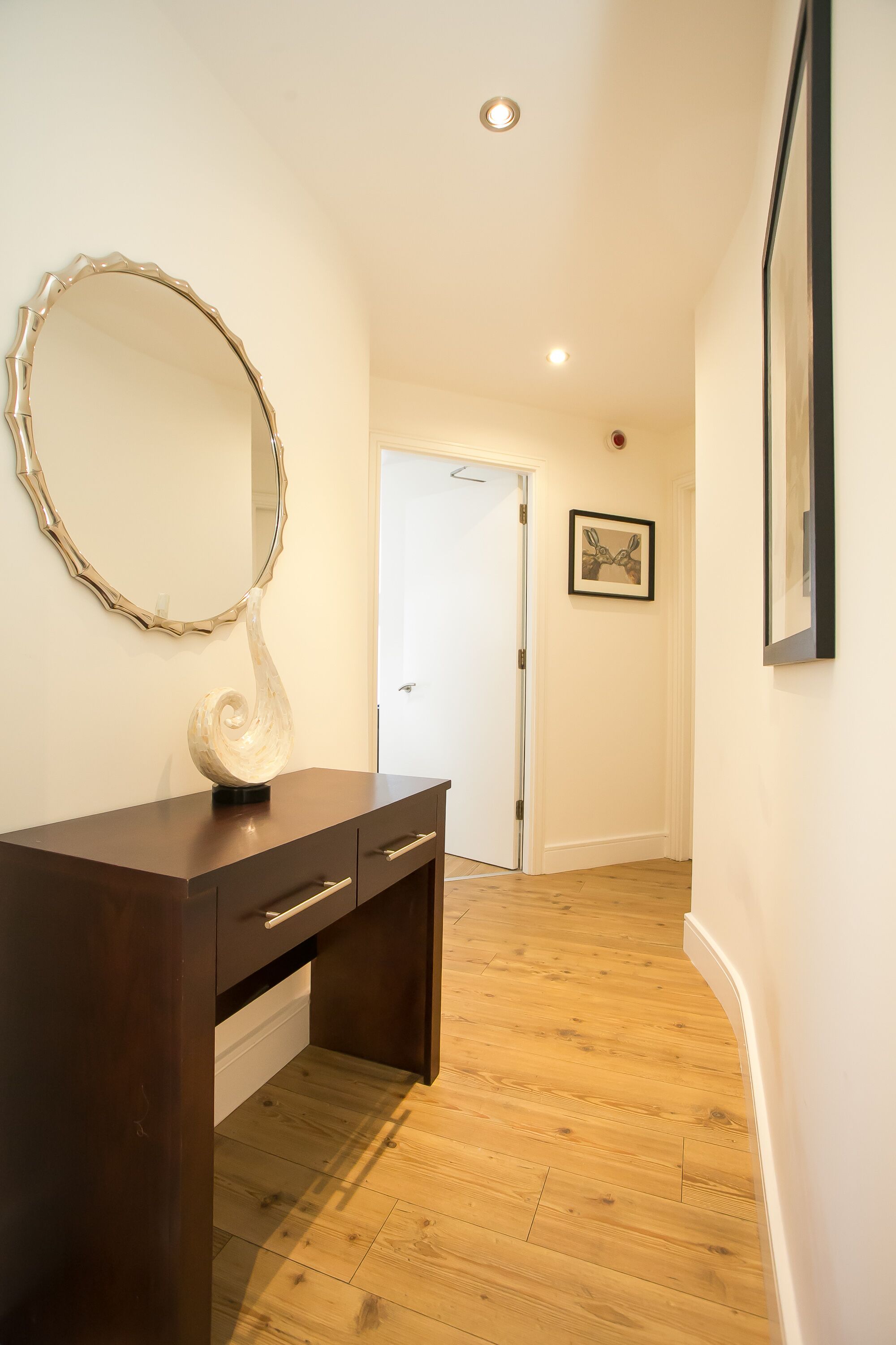 A hallway with a wooden floor, a dark wooden dresser with drawers, a large mirror, and a framed picture on the wall.