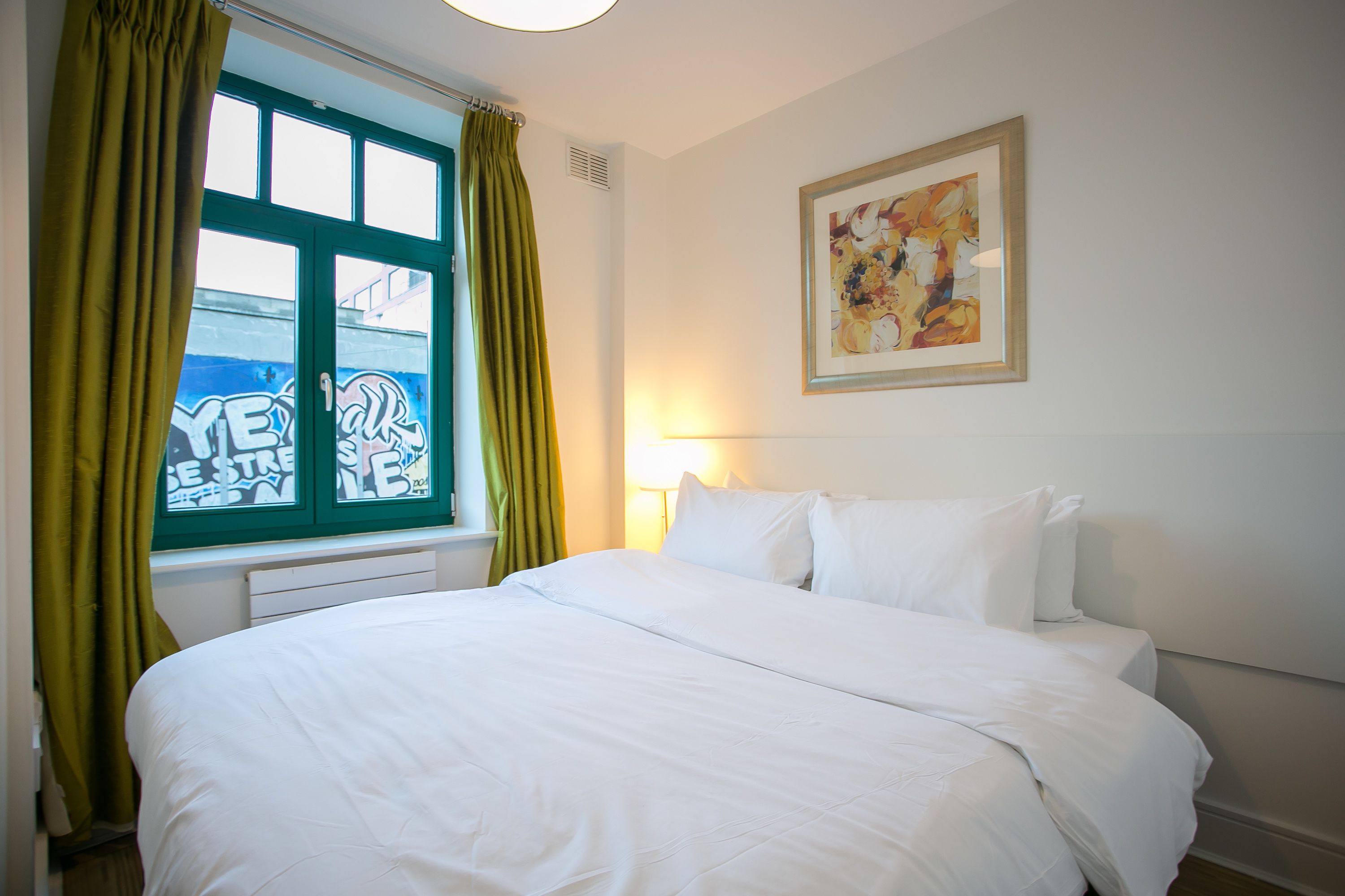 A hotel room with a bed, a window with green curtains, a wall-mounted painting, and a bedside lamp.