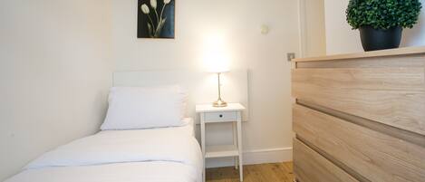 A bedroom with a bed, a nightstand with a lamp, a wall-mounted painting, and a potted plant.