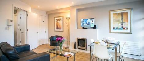 A modern living room with a black leather sofa, a glass coffee table, a fireplace, a TV, and a dining area with white chairs.