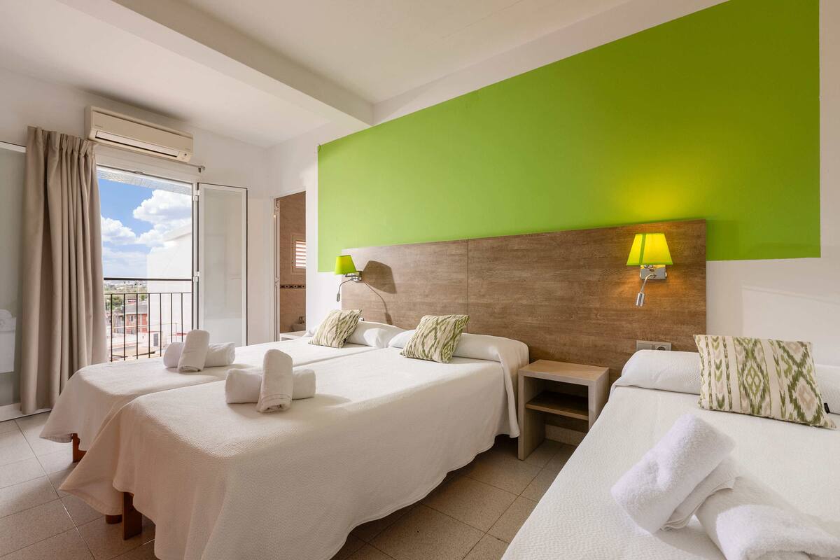 Ein Hotelzimmer mit zwei Betten, einer grünen Akzentwand, einem Balkon mit Vorhängen und einem Nachttisch mit Lampe.