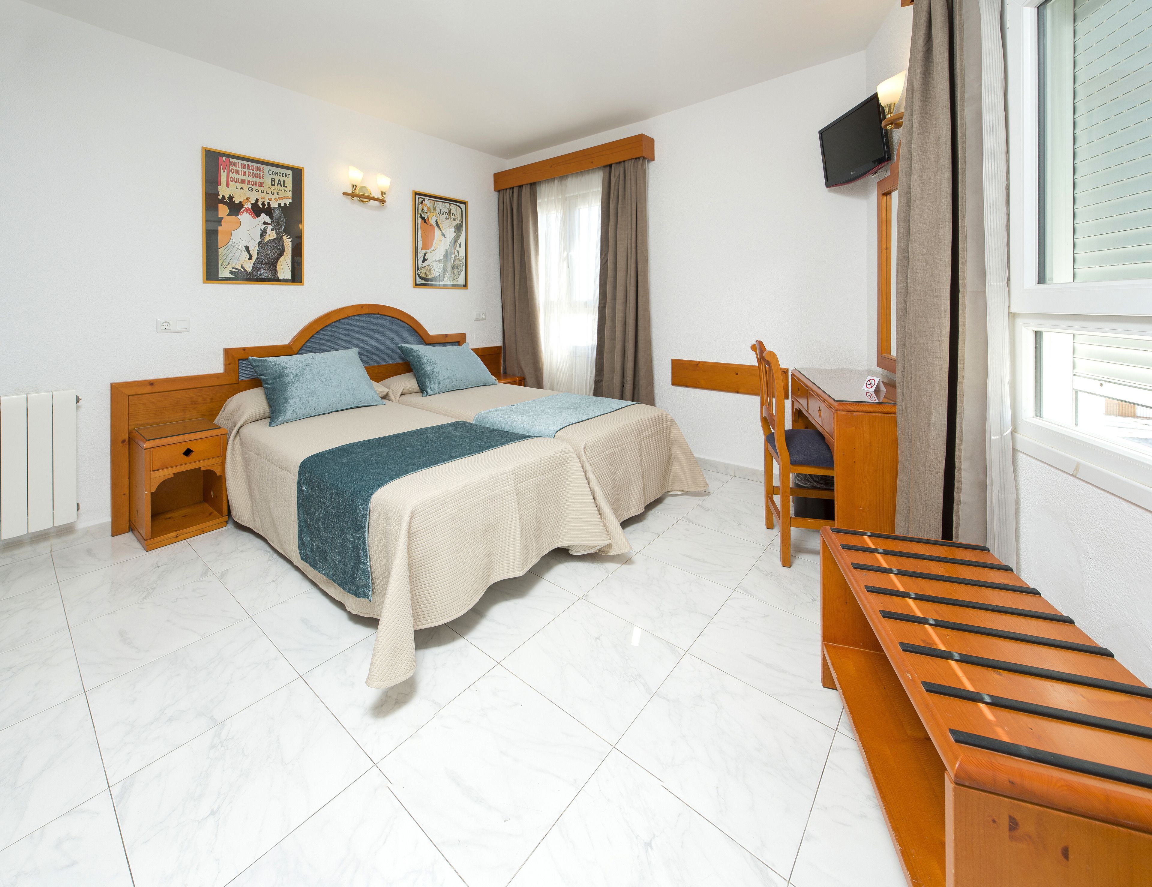 A hotel room with a bed, bedside tables, a desk, a chair, a television, and a window with curtains.