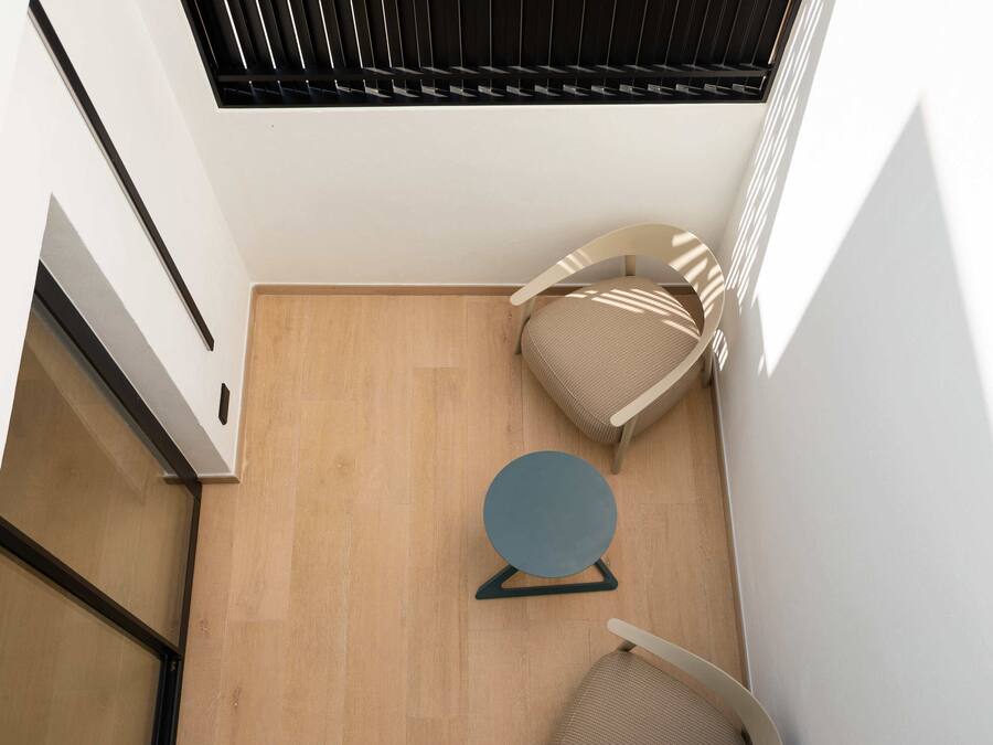 Una habitación moderna con piso de madera, dos sillas color beige, una mesita redonda y una ventana con persianas.