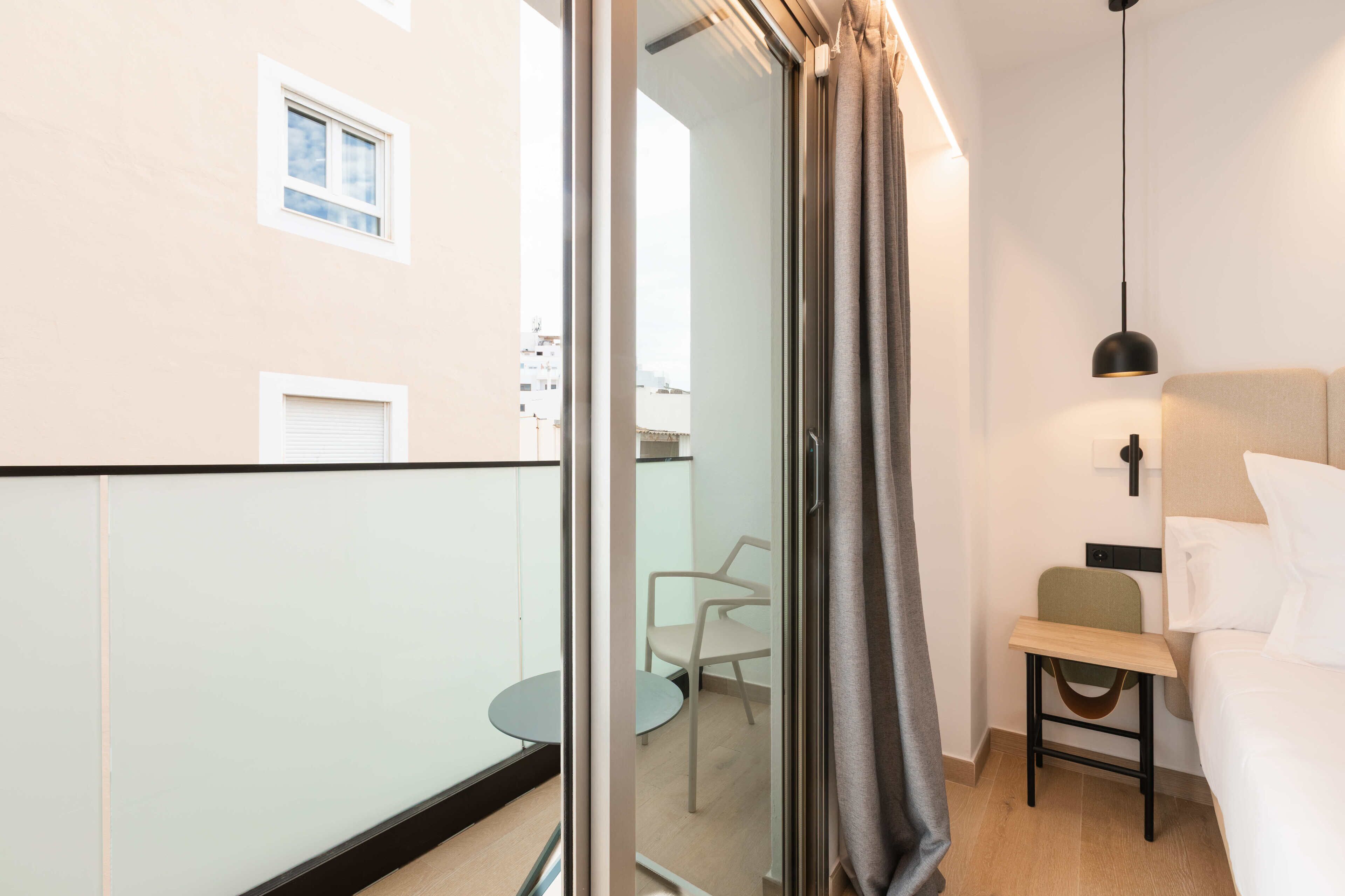 A modern hotel room with a bed, a small desk, a chair, and a view of the city through the window.
