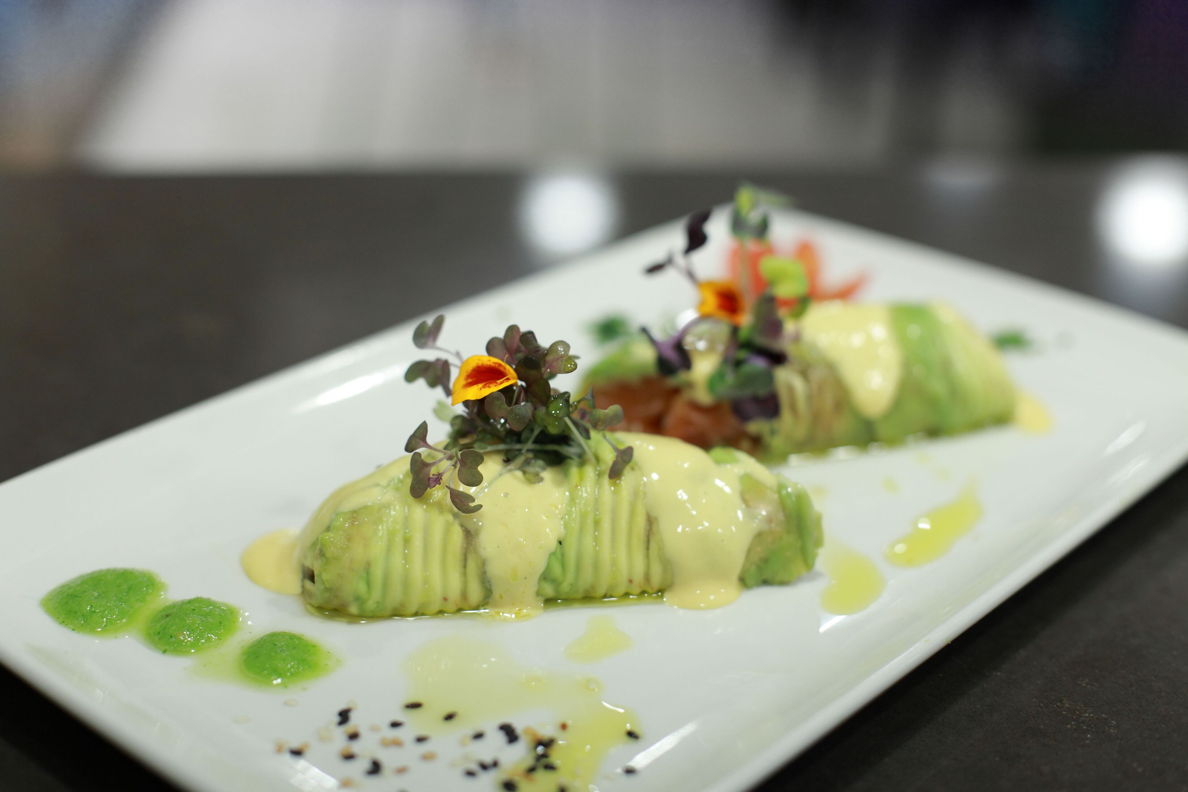 Un plato rectangular blanco con un guiso de verdura verde, posiblemente calabacín relleno, cubierto con salsa cremosa y decorado con brotes tiernos y una pequeña flor naranja.