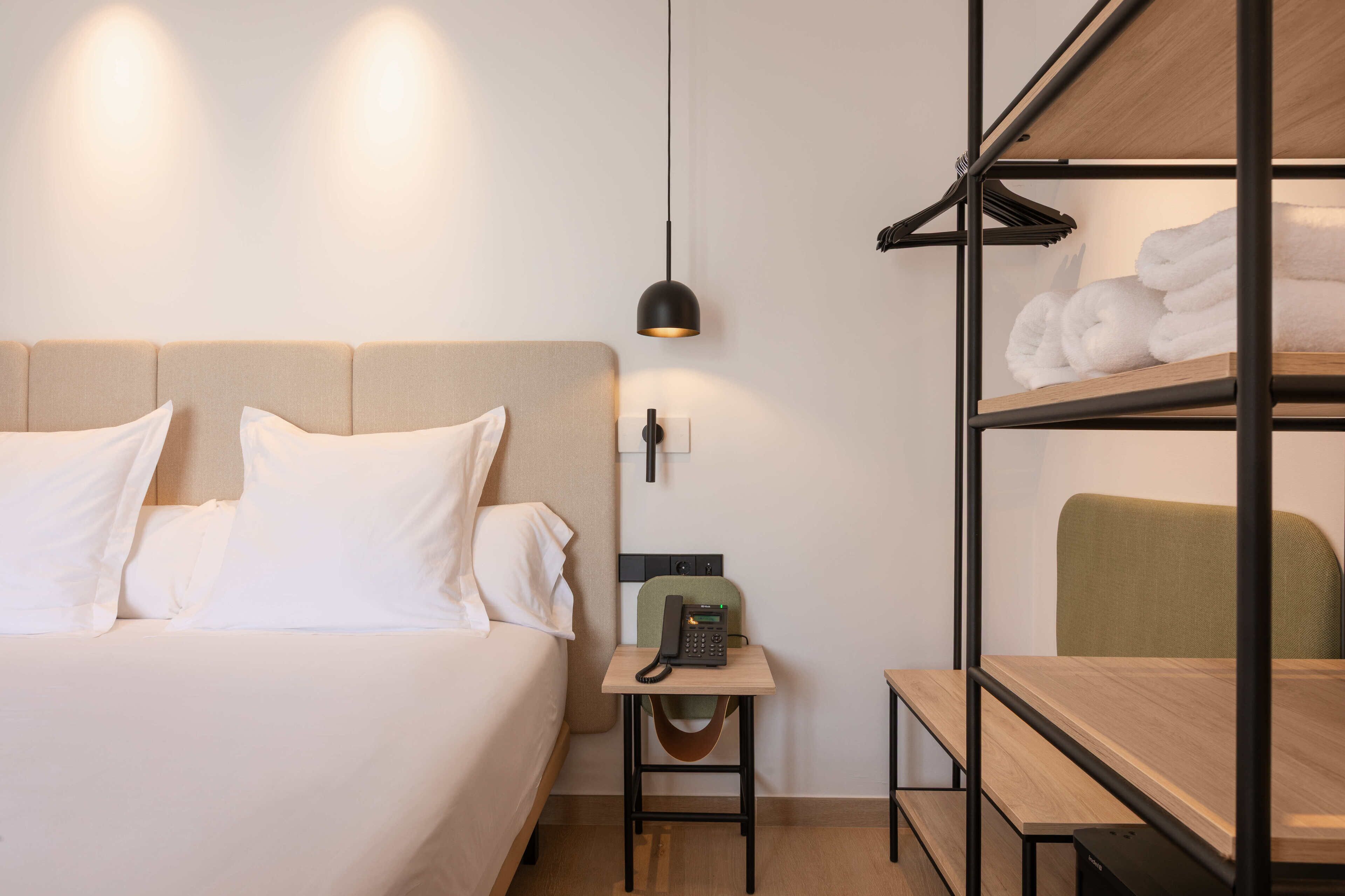 Una habitación de hotel con cama, mesita de noche con teléfono, lámpara colgante, estante con toallas y una silla.