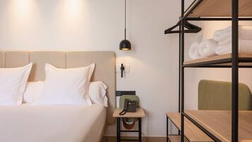 A hotel room with a bed, bedside table with a phone, a hanging light, a shelf with towels, and a chair.