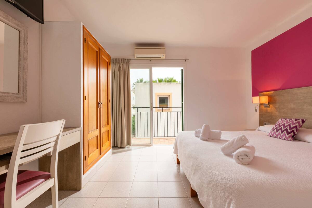 Ein Hotelzimmer mit Bett, Schreibtisch, Stuhl, Kleiderschrank und einem Balkon mit Blick auf Gebäude.