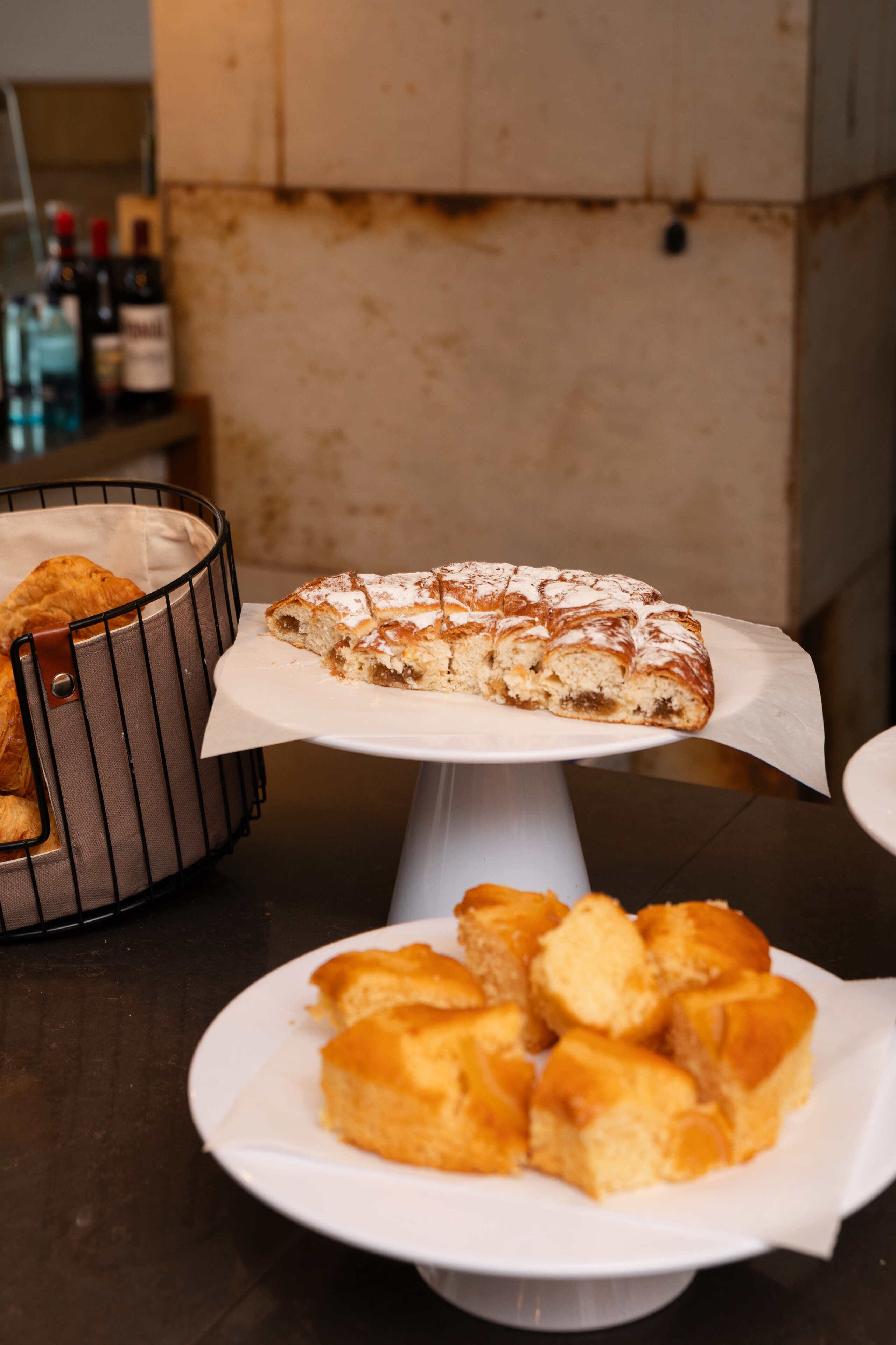 Un présentoir à desserts avec une part de gâteau sur un présentoir blanc, un panier rempli de viennoiseries et une assiette contenant plusieurs morceaux de gâteau.