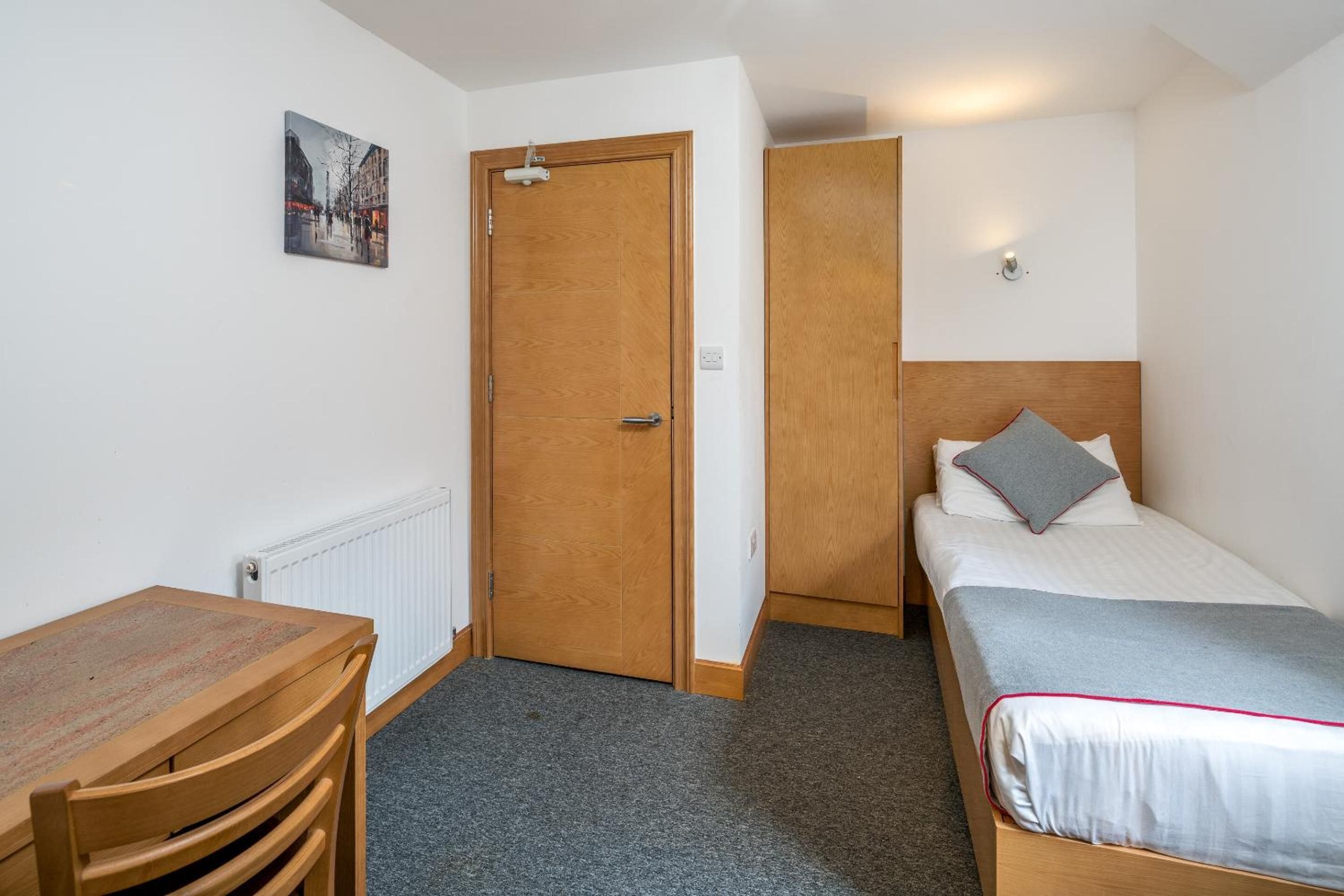 A small, clean room with a wooden desk, a bed with a grey pillow, and a door leading to another room.