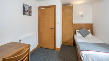 A small, clean room with a wooden desk, a bed with a grey pillow, and a door leading to another room.