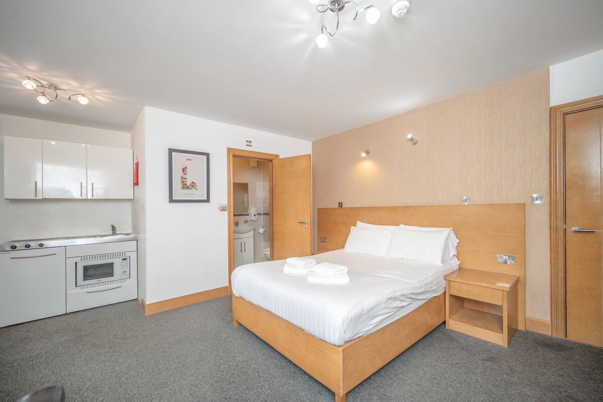 A hotel room with a bed, a wooden headboard, a small bedside table, a built-in microwave, and a kitchenette with cabinets and a sink.