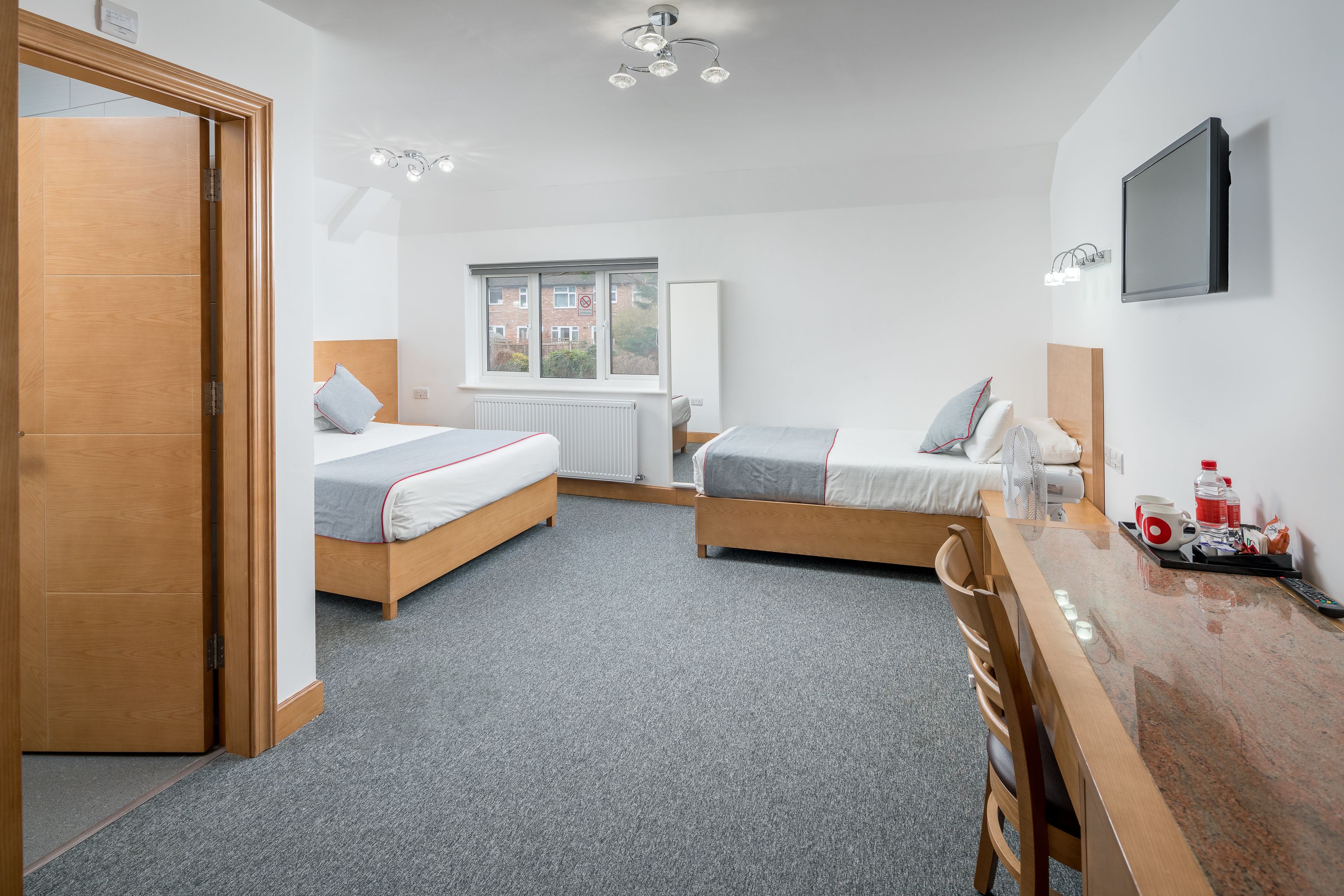 A modern hotel room with two single beds, a wooden desk, a chair, a TV mounted on the wall, and a window with a view of buildings outside.
