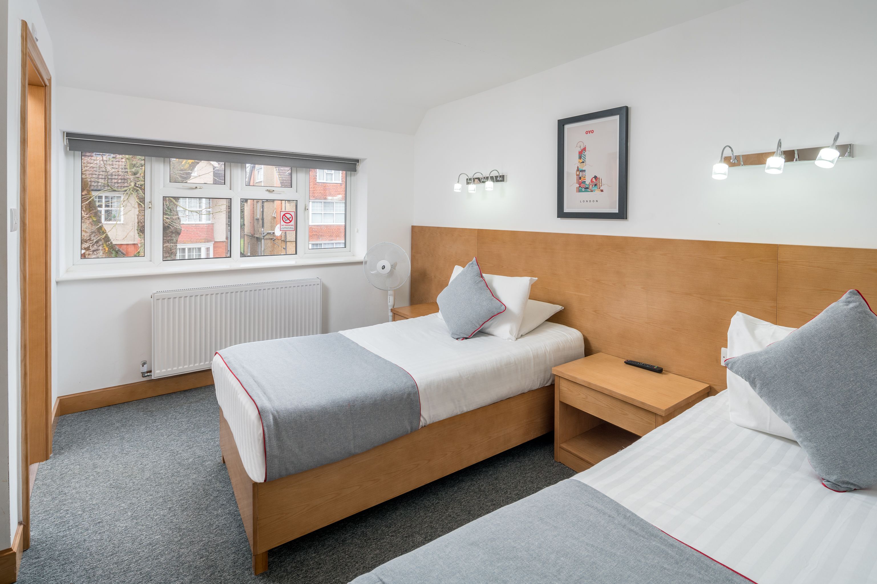 Moderni hotellihuone, jossa on suuri sänky, yöpöytä, pieni työpöytä, ikkuna, jossa on sälekaihtimet, patteri ja seinällä oleva kehystetty taulu.