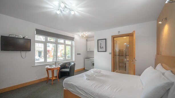 A hotel room with a bed, a TV mounted on the wall, a small table with a chair, a window, and a kitchenette. - Flexistay Aparthotel Sutton (Sutton)