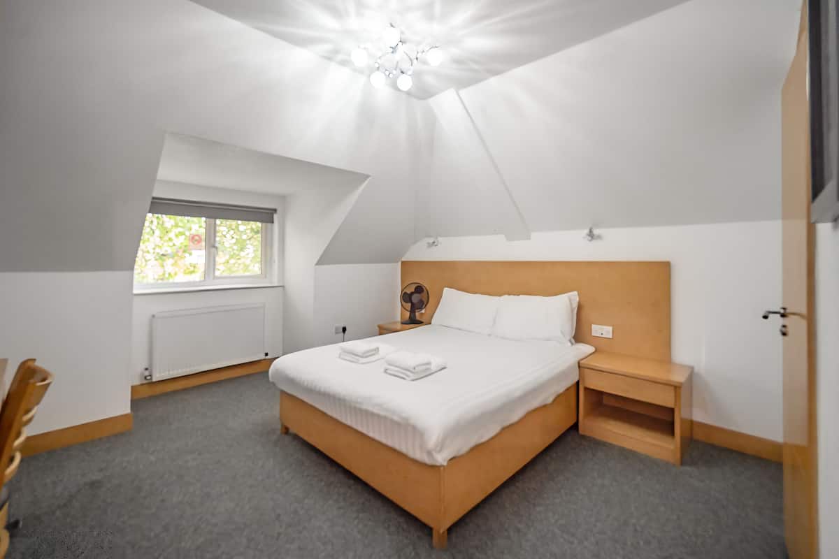 A bedroom with a large bed, a nightstand, a wooden headboard, a window with blinds, and a ceiling fan.
