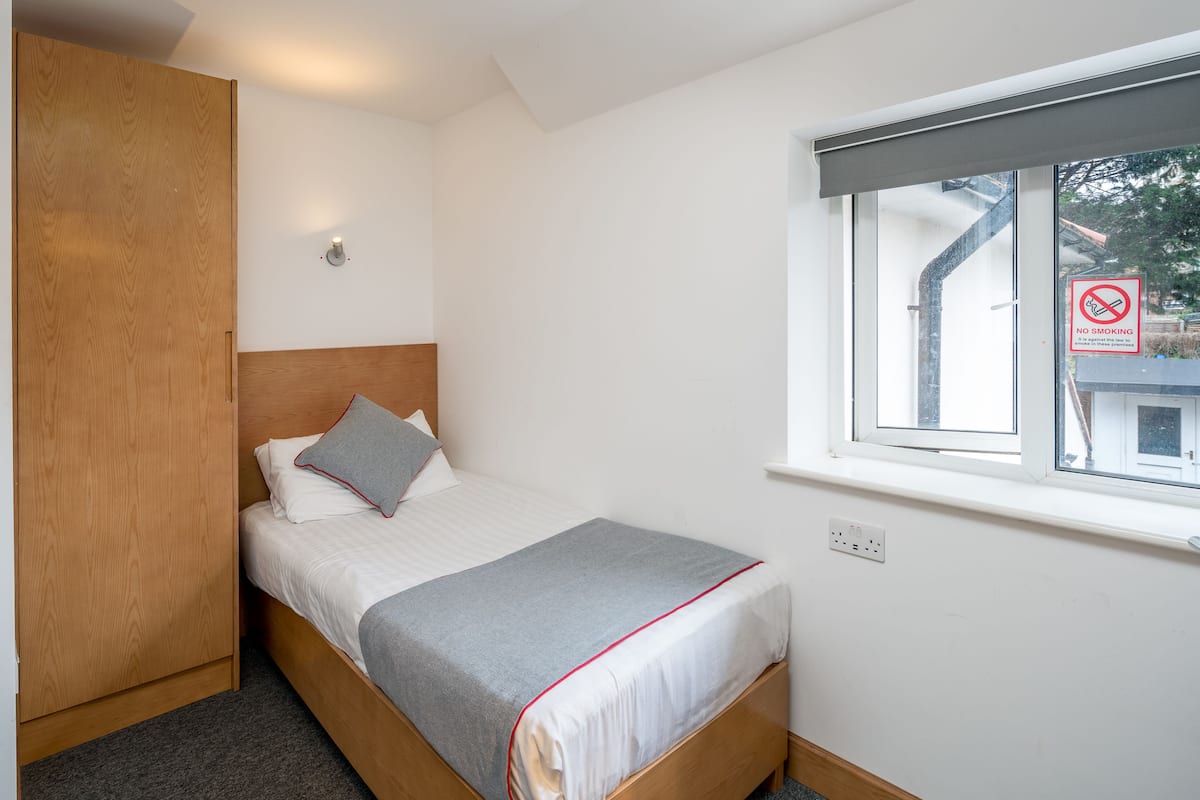 A small, neatly arranged bedroom with a single bed, a wooden wardrobe, a window with a view, and a 'No Smoking' sign.