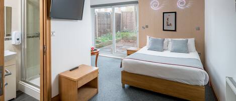 Habitación de hotel con cama, televisión montada en la pared, una pequeña mesa de madera y zona de ducha.