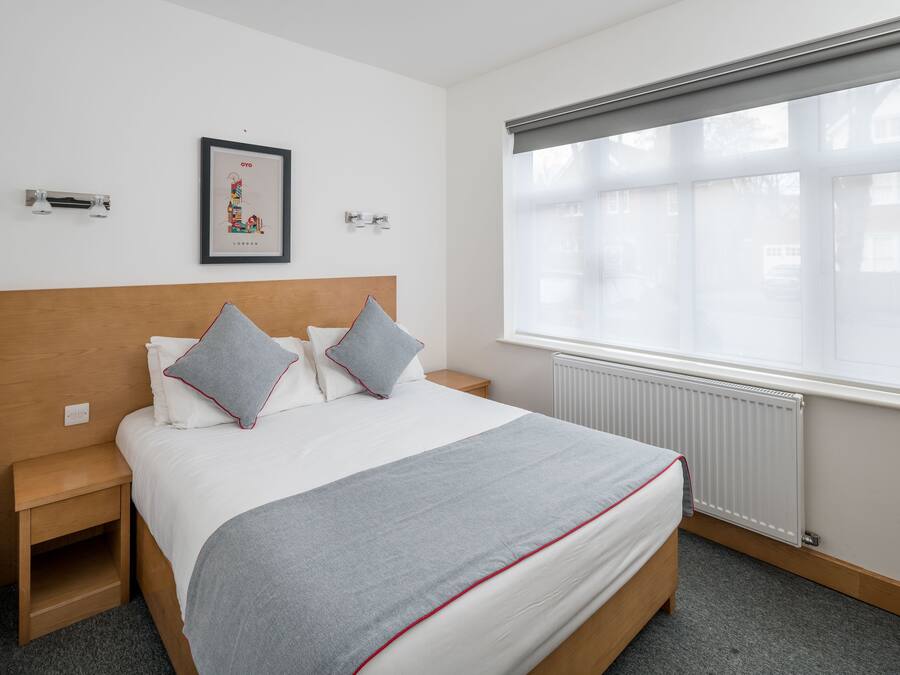 Habitación de hotel ordenada con una cama grande, una mesita de noche, un cuadro enmarcado en la pared y una ventana con persianas.
