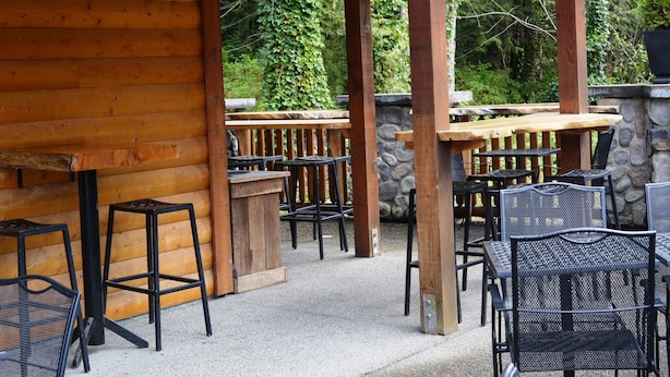 Eine Terrasse im Freien mit Holztischen und Metallstühlen, umgeben von Steinmauern und Grünpflanzen.
