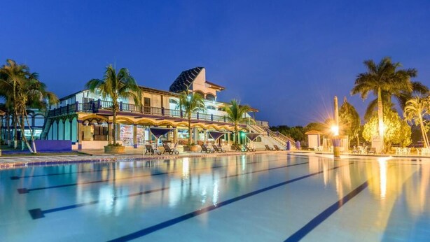 Ein großer Pool mit Liegestühlen und Palmen, umgeben von einem Gebäude mit mehreren Balkonen und einem überdachten Bereich.