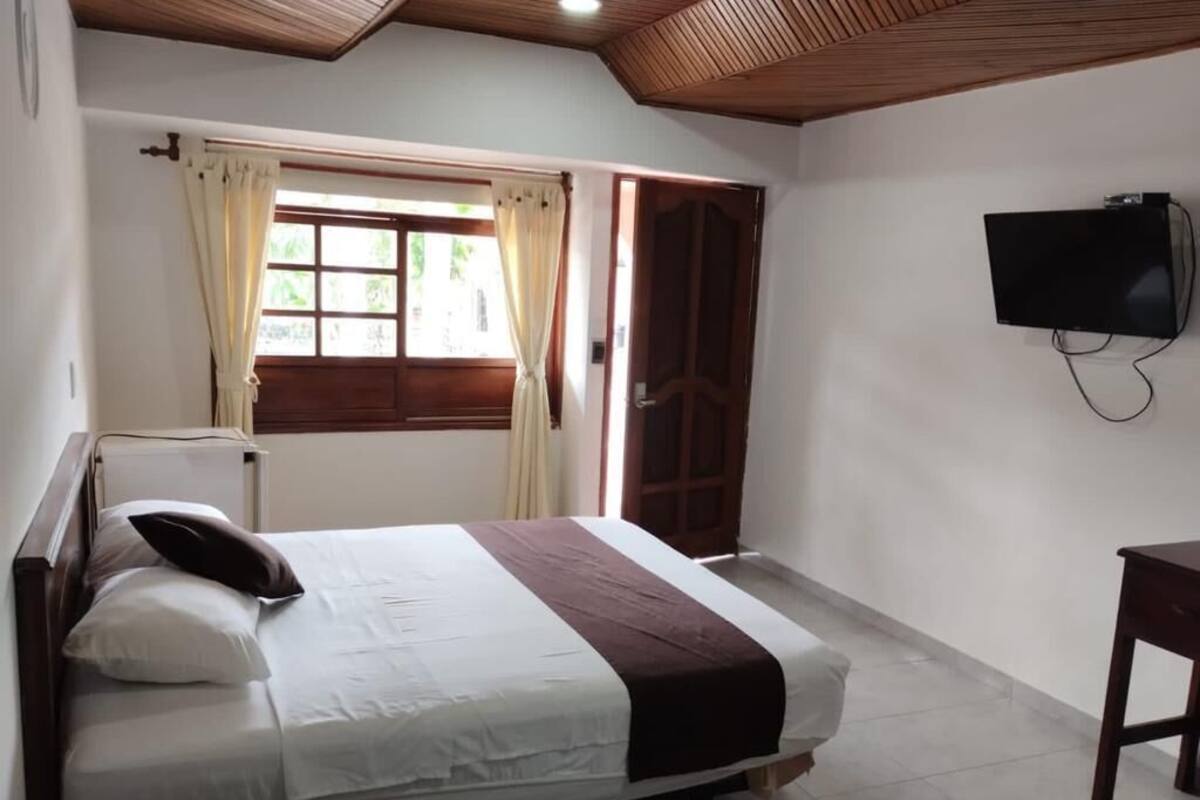Une chambre à coucher avec un lit, un téléviseur fixé au mur, une fenêtre avec des rideaux et une porte en bois.