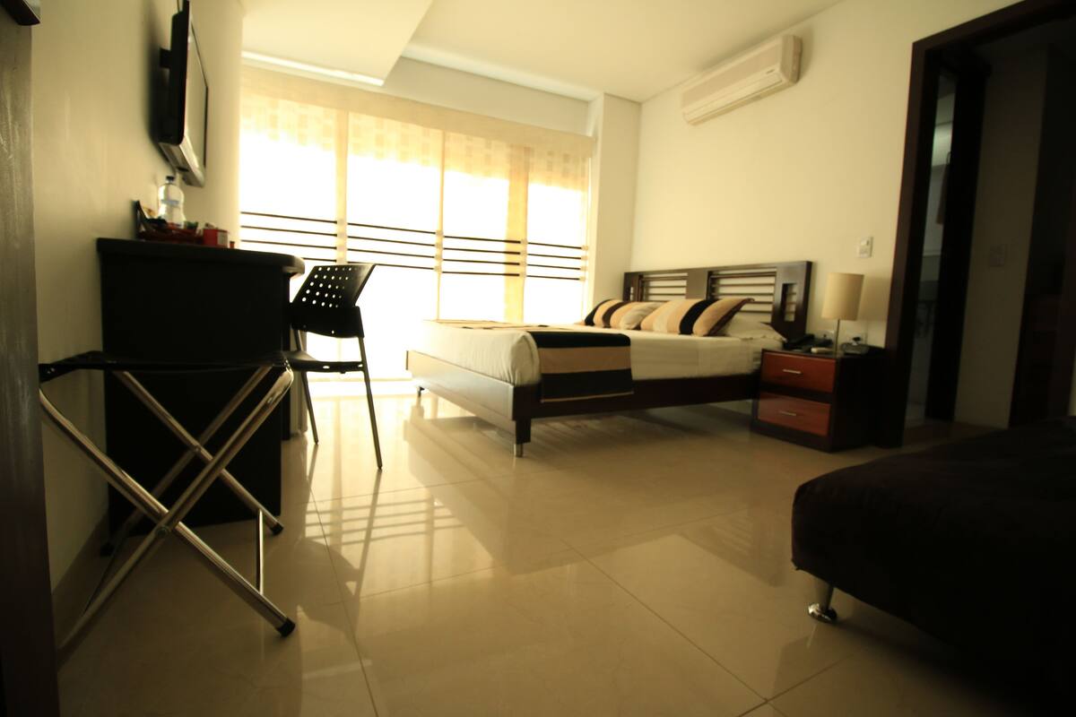 A hotel room with a bed, a desk, a chair, a nightstand, and a window with curtains.