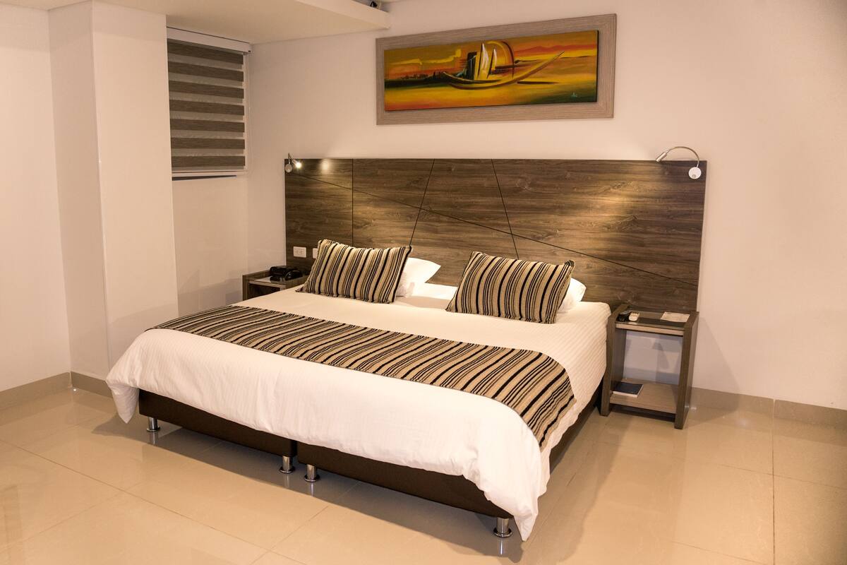 A modern bedroom with a large bed, striped bedding, a wooden headboard, a nightstand, and a painting on the wall.