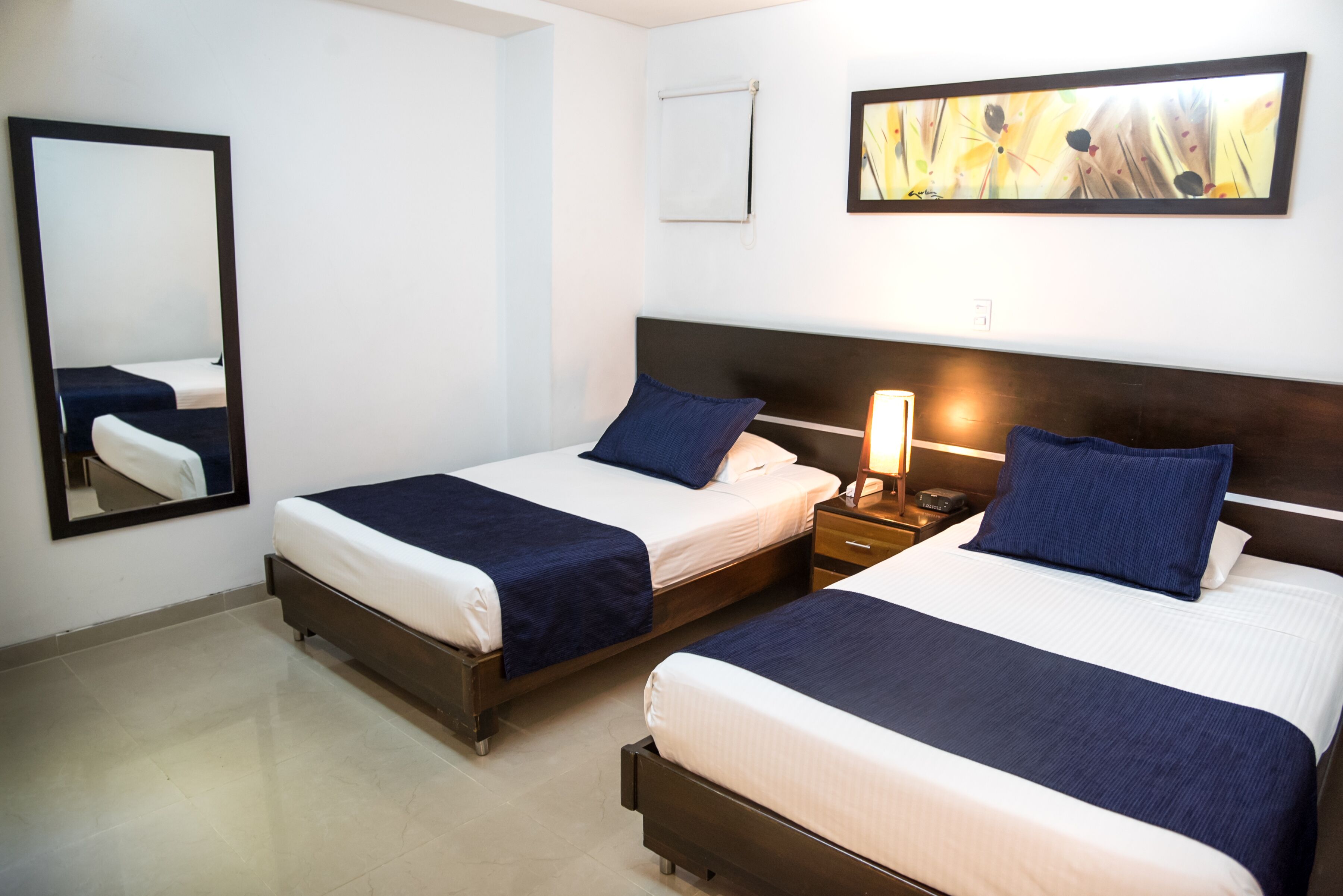 Quarto de hotel com duas camas, uma mesa de cabeceira com abajur, um espelho e um quadro na parede.