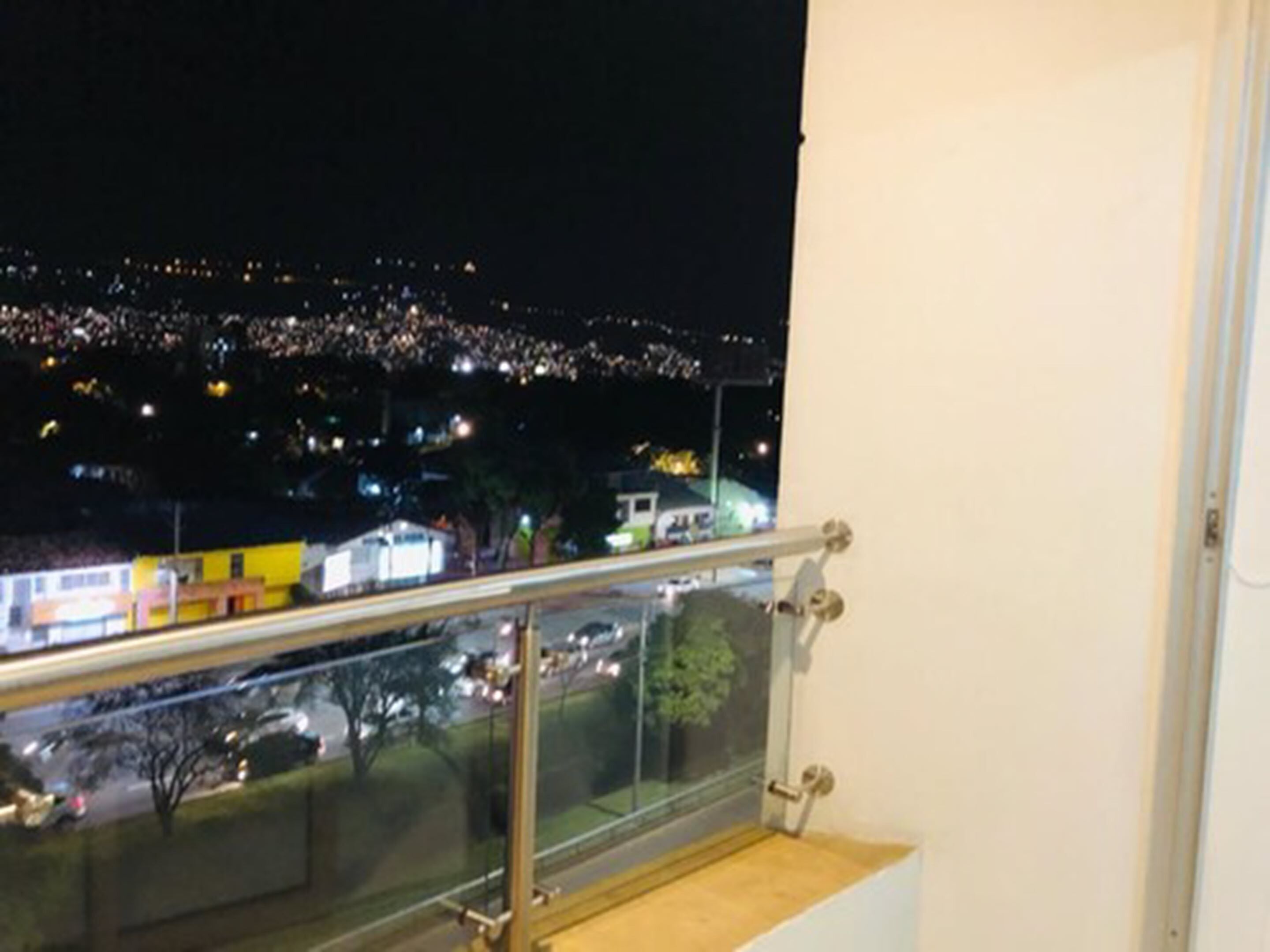 A night view from a balcony with a railing, overlooking a cityscape with illuminated buildings and streets.