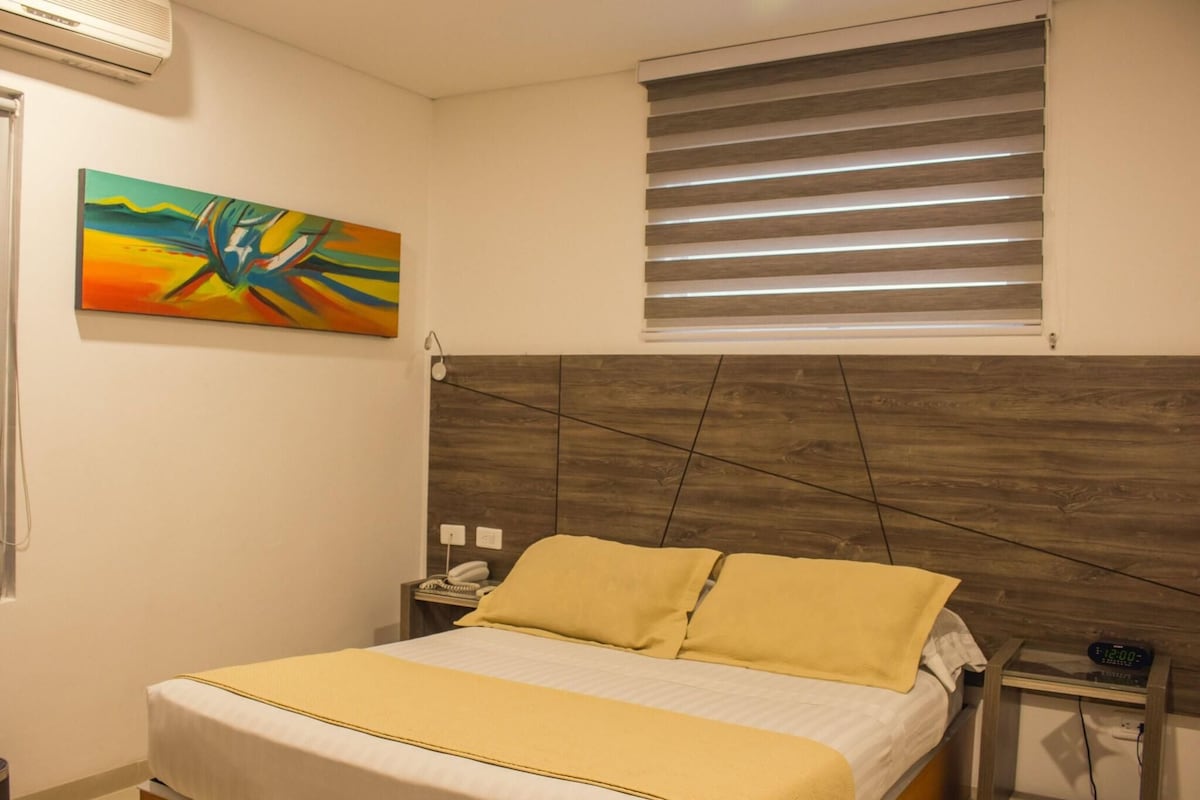 Habitación de hotel con cama, un cuadro, una mesita de noche, ventana con persianas y aire acondicionado instalado en la pared.