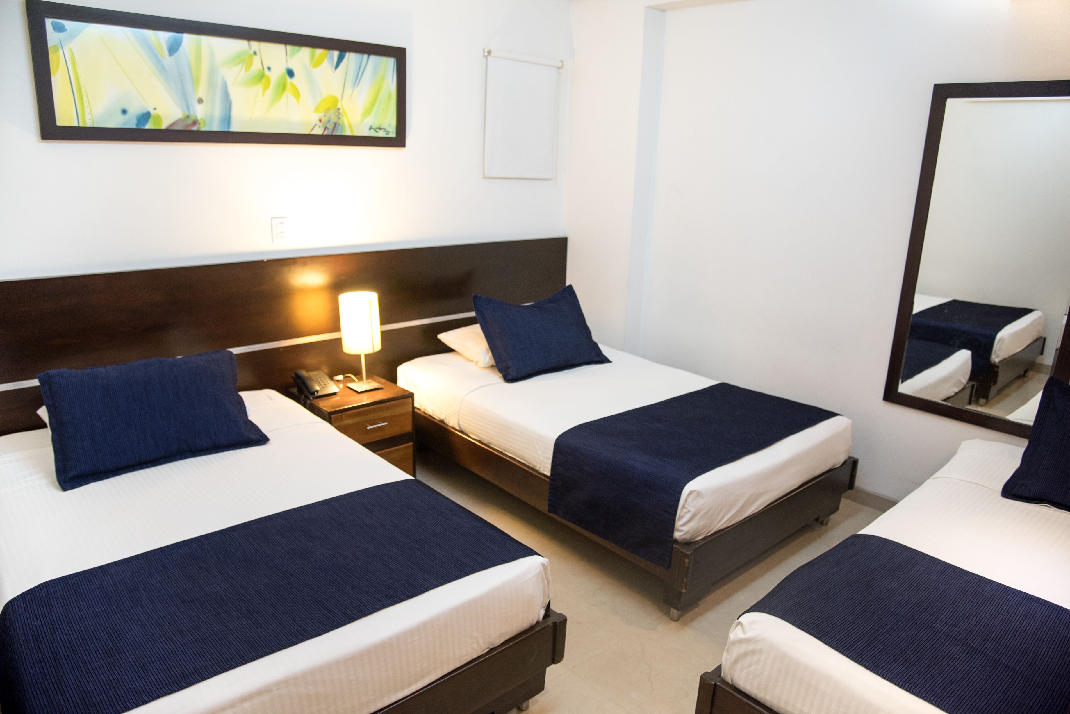 Une chambre d’hôtel avec deux lits, chacun recouvert d’une couvre-lit bleue et doté de draps blancs. On y trouve une table de chevet avec une lampe, une œuvre d’art accrochée au mur et un miroir.