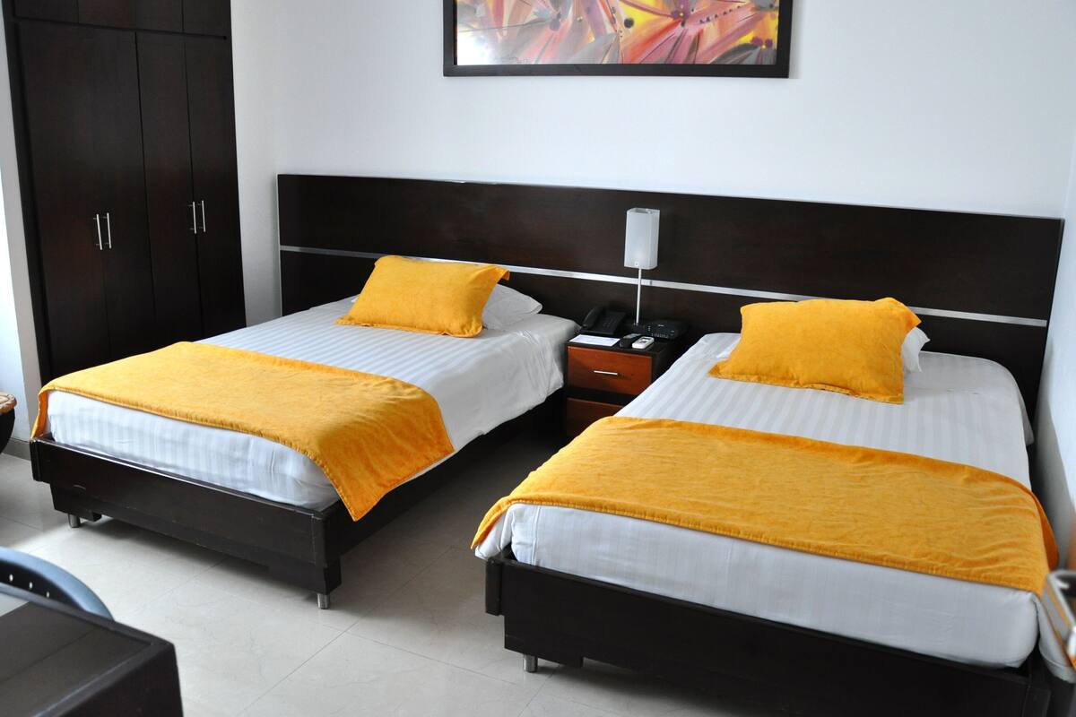 Two single beds with white bedding and orange throw pillows, a nightstand with a phone and lamp, and a dark wardrobe.