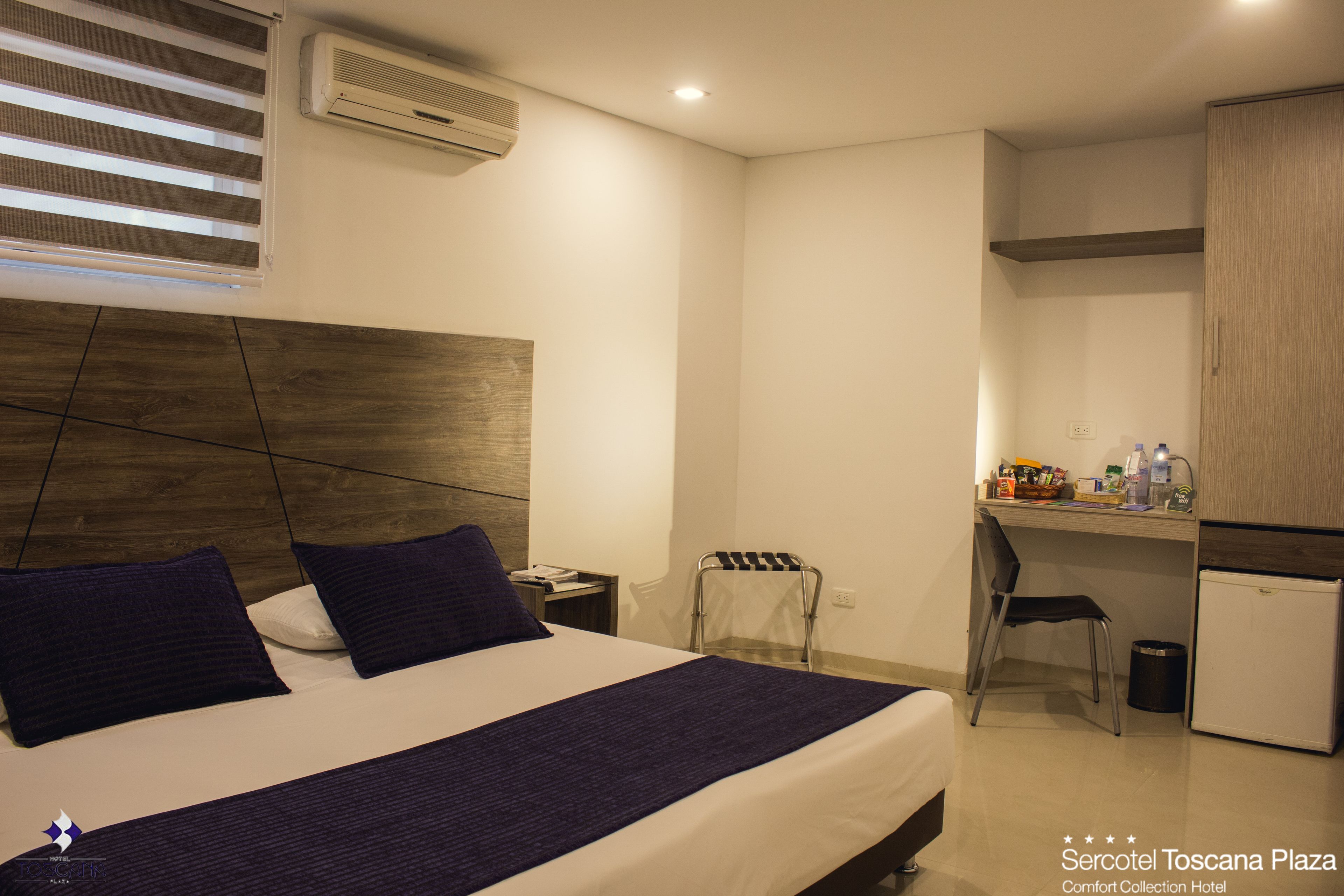 A hotel room with a bed, a desk, a chair, and a small refrigerator.