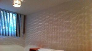 Un dormitorio con colcha estampada, ventana con persianas, pared con textura y una mesita de noche roja.