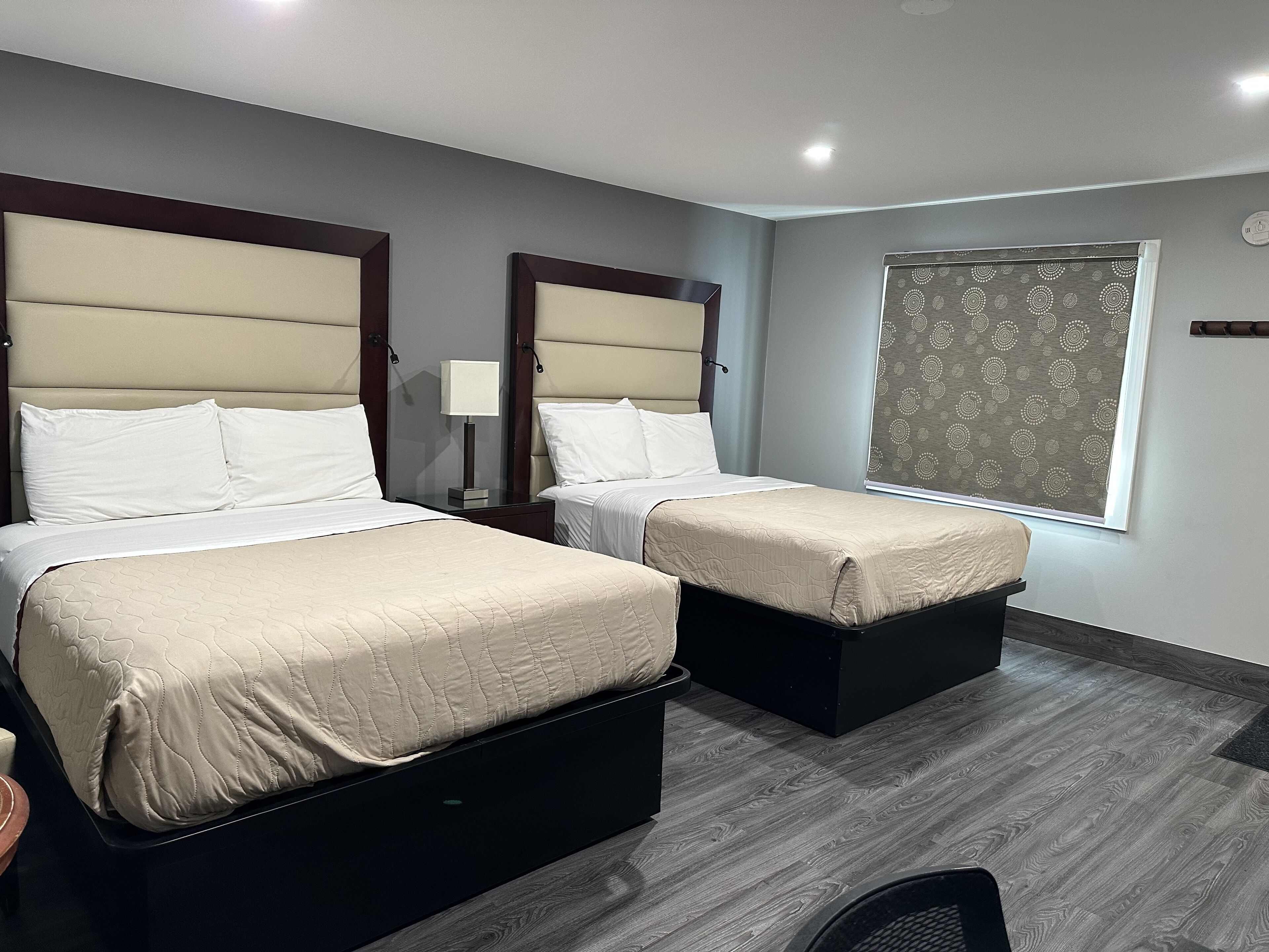 A hotel room with two beds, a bedside table with a lamp, a window with patterned curtains, and a grey wooden floor.
