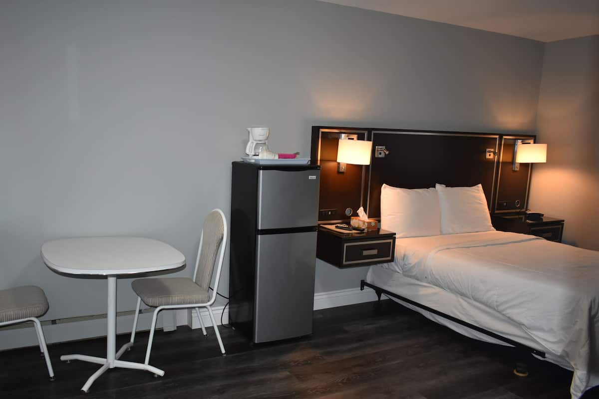 A hotel room with a bed, a small table with two chairs, a refrigerator, and a nightstand with a lamp.