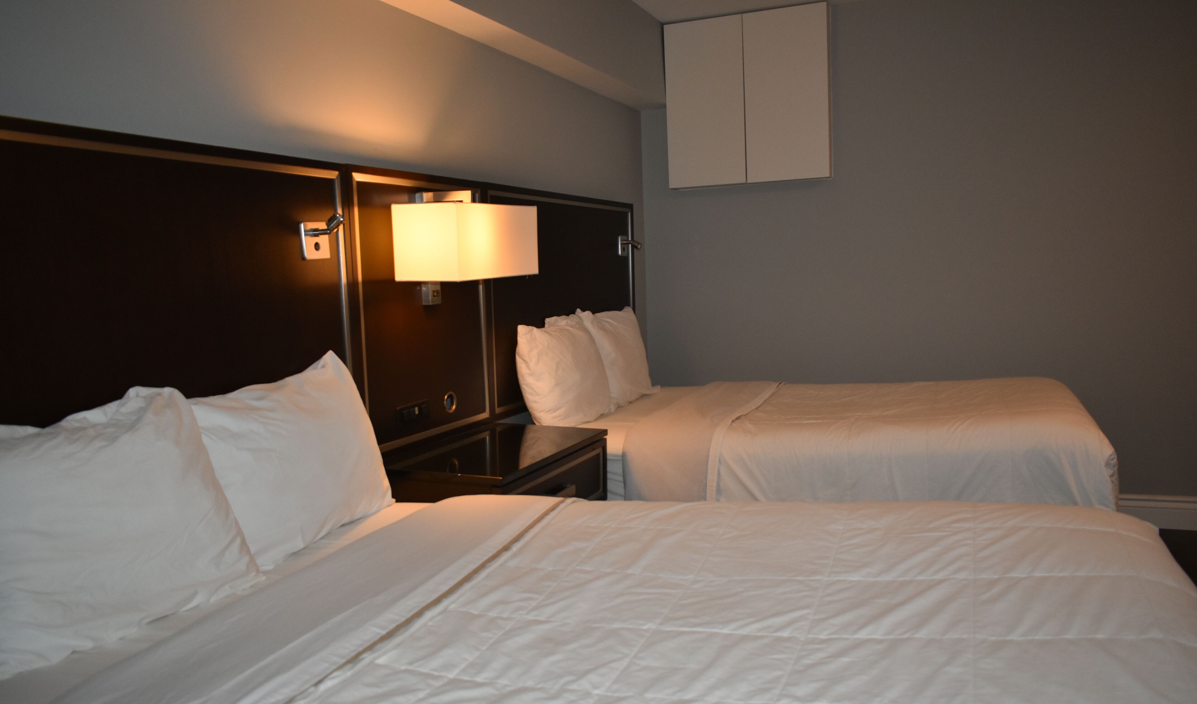 A hotel room with two beds, a nightstand, a lamp, and a wall-mounted cabinet.