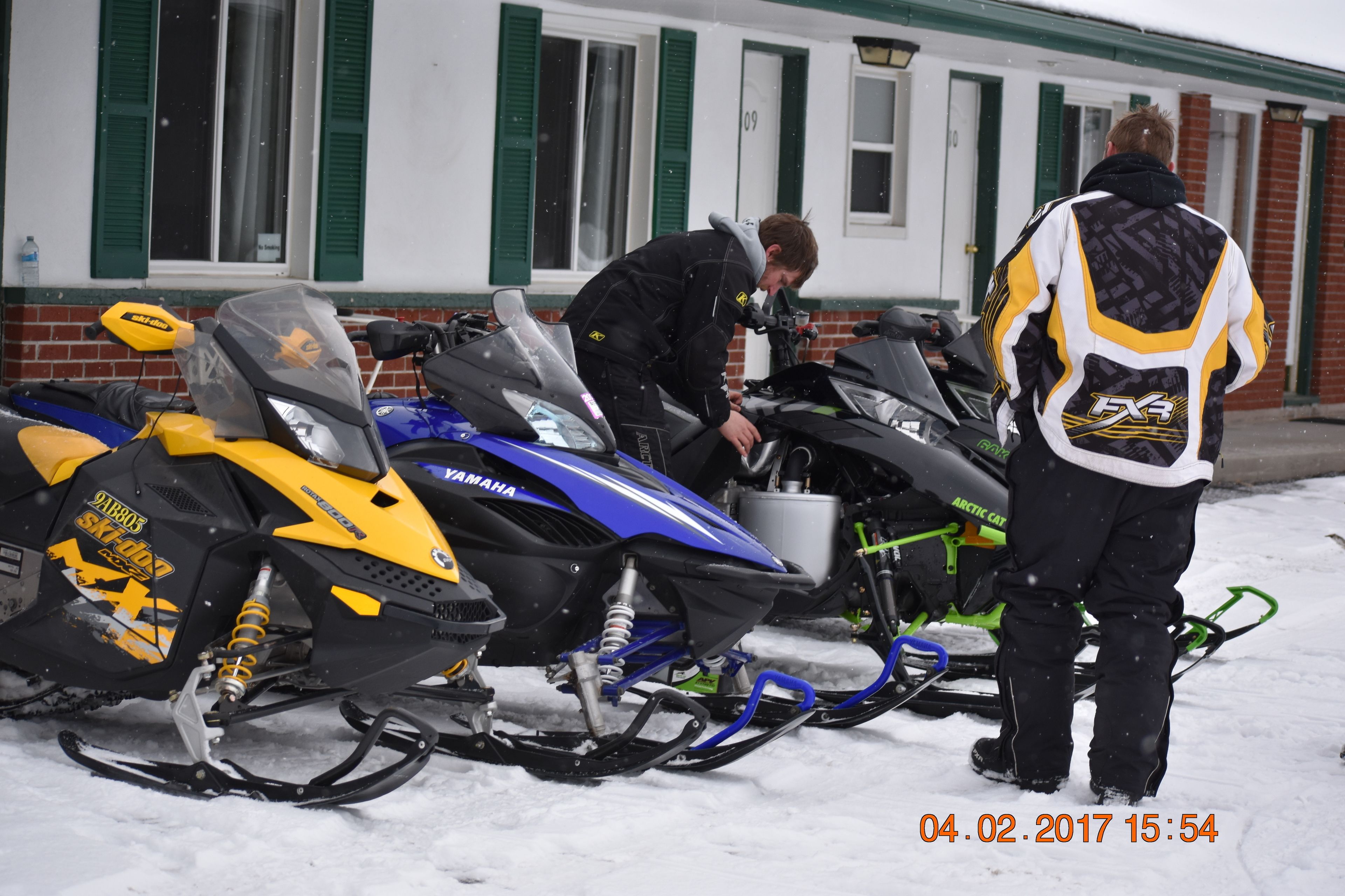 Three snowmobiles parked in front of a building with green shutters.