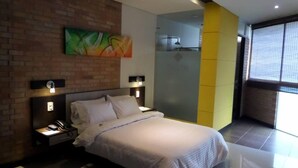 A modern bedroom with a brick accent wall, a large bed, two bedside tables with lamps, a painting, and a large window. - Hotel Toscana Oriente (El Carmen de Viboral)