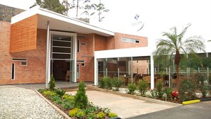 A modern building with a brick facade, large glass windows, and a landscaped garden area with various plants and a palm tree. - Hotel Toscana Oriente (El Carmen de Viboral)