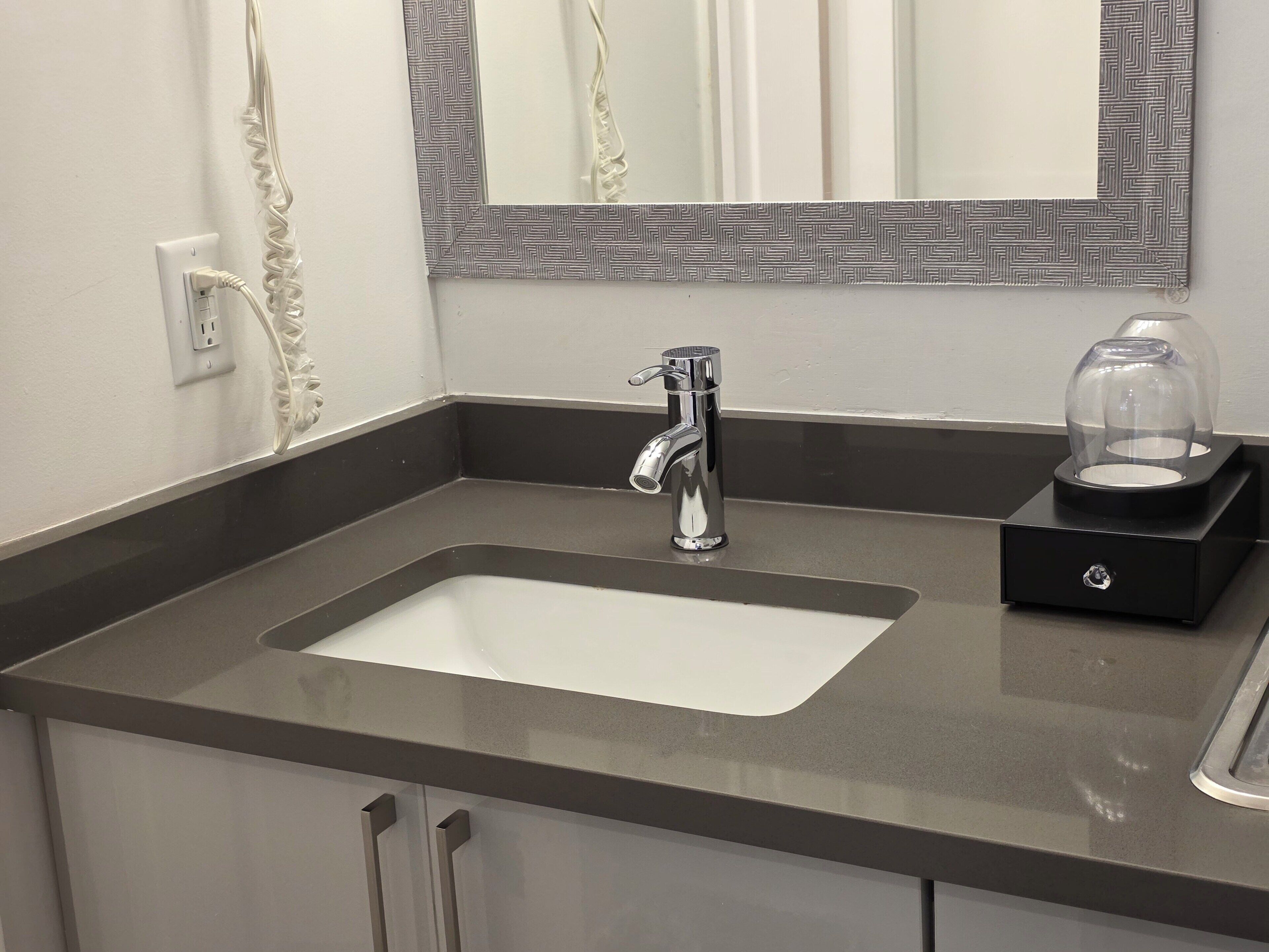 A modern bathroom with a chrome faucet, a mirror with a patterned frame, and a glass-enclosed soap dispenser.