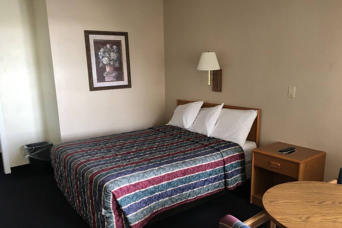 Hotellihuone, jossa on sänky, yöpöytä lampun kanssa, tuoli ja pieni pyöreä pöytä.