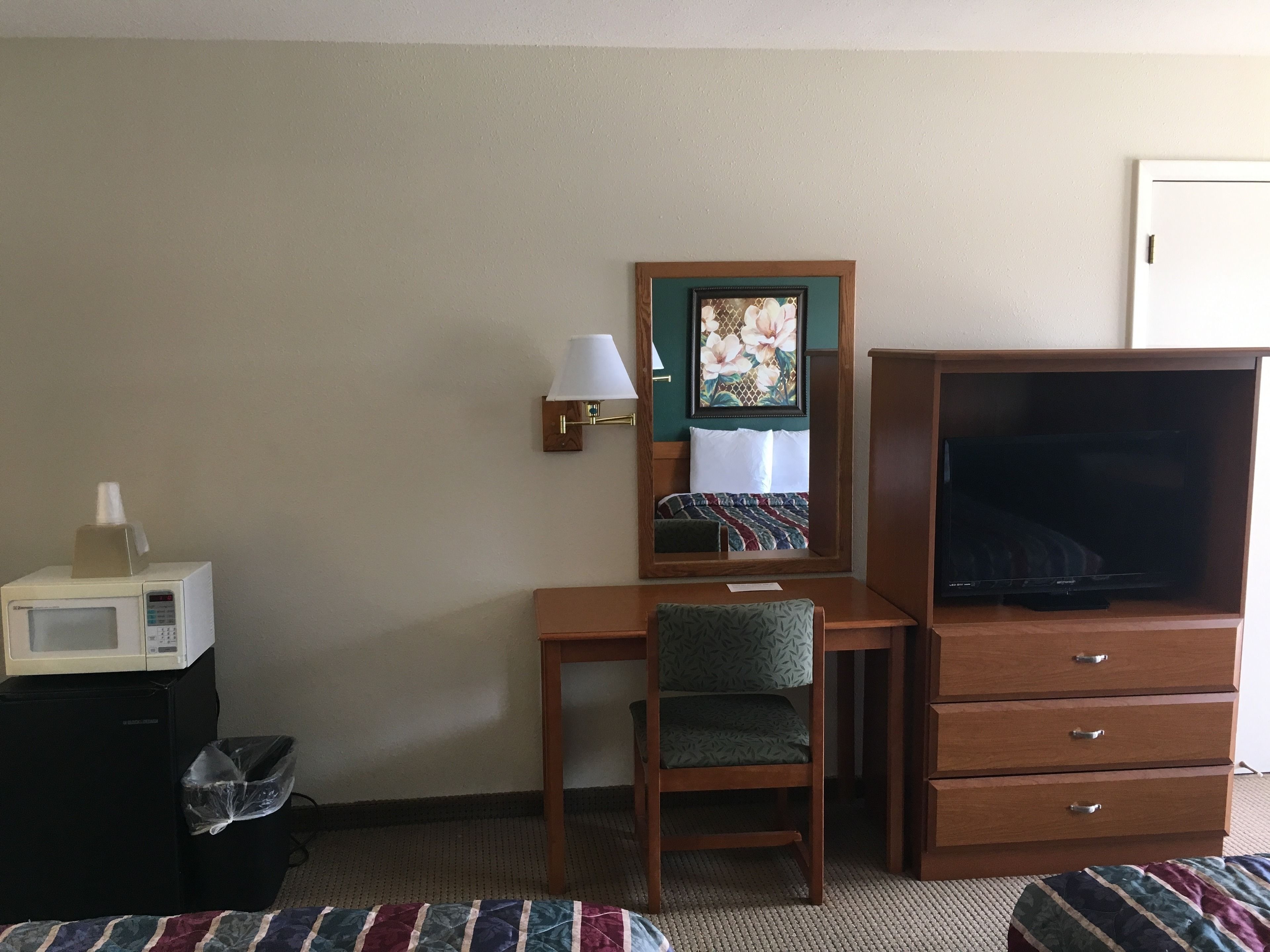 A hotel room with a wooden desk, a chair, a television, a microwave, and a trash bin.