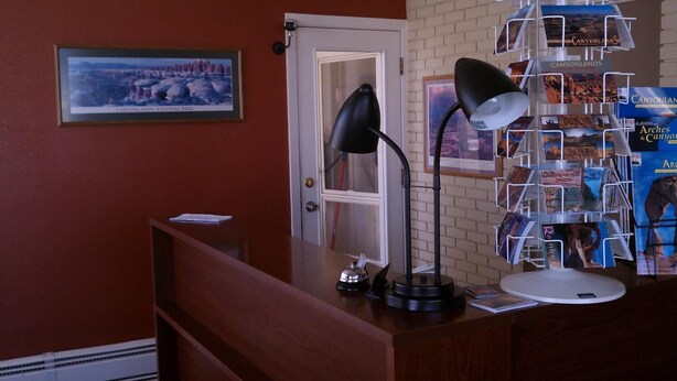 A reception desk with a framed picture of Canyonlands National Park, a black desk lamp, a white desk lamp, and a display rack with travel brochures. - The Monticello Inn (Monticello)