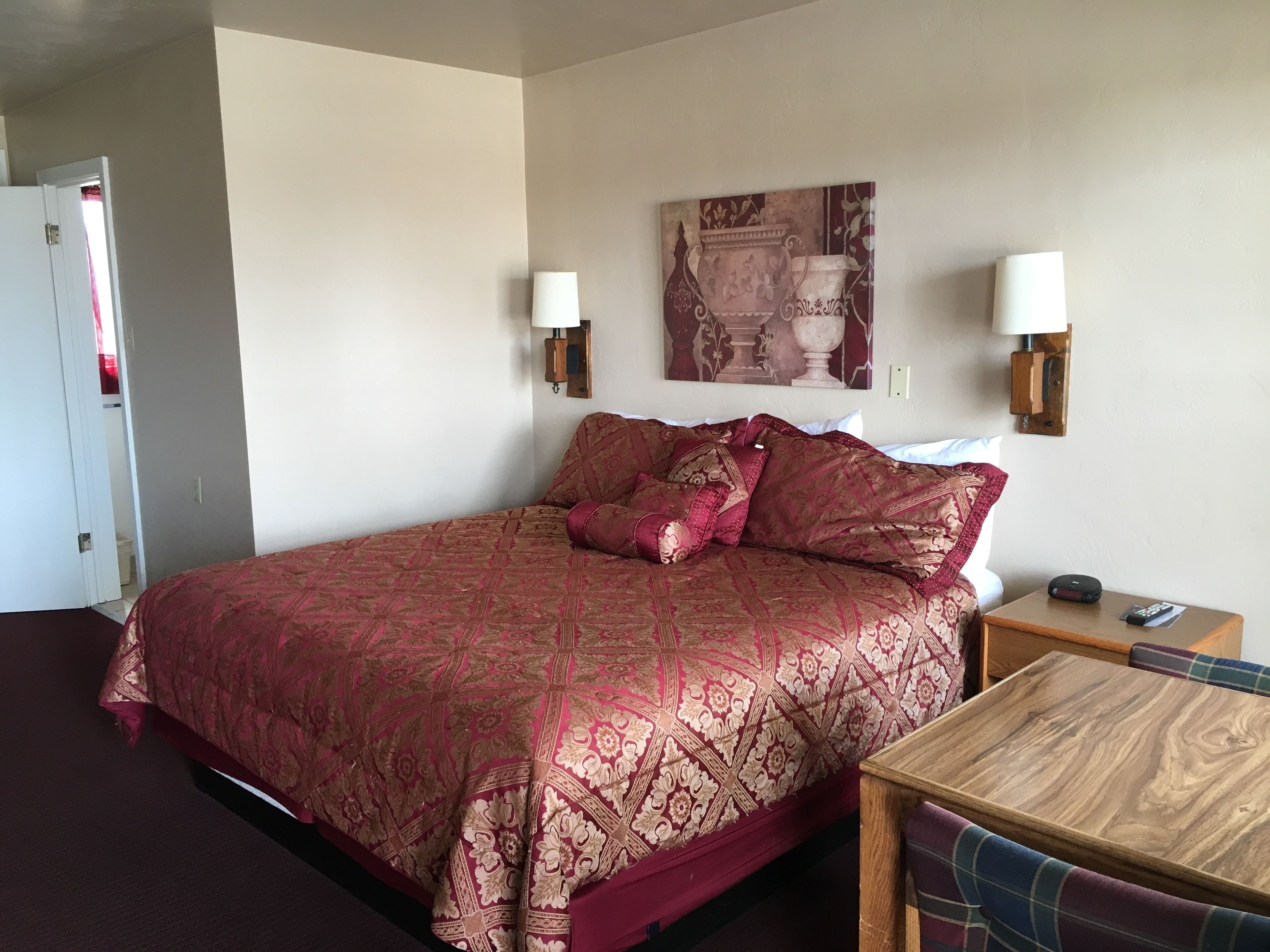 A hotel room with a large bed, a wooden nightstand, a small table, and a painting on the wall.