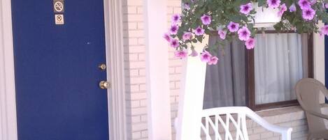 Una puerta azul con una placa numérica, un banco blanco y una cesta de flores colgante.